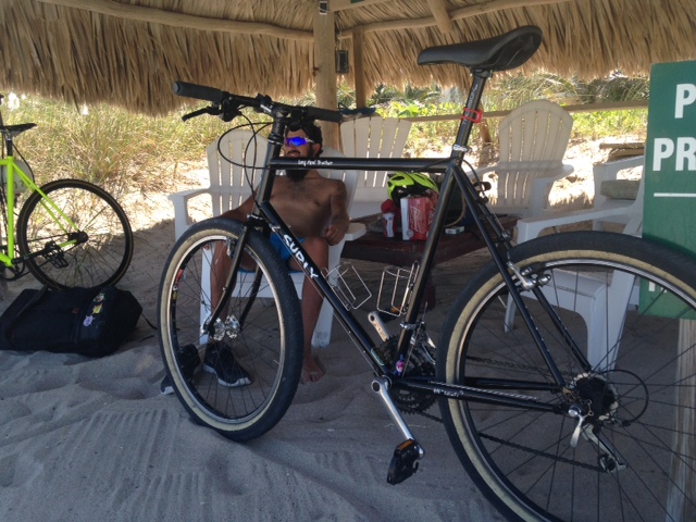 Left side view of a black Surly Long Haul Trucker bike, with a person sitting behind, on sand under a grass roof shelter