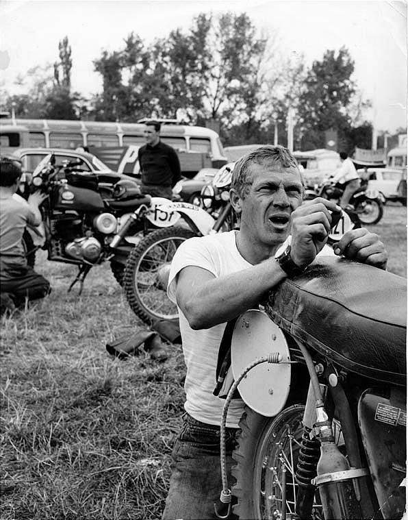 Steve McQueen kneeling in grass behind a motorcycle - black and white image