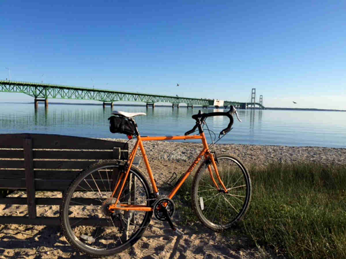Right side view of an orange Surly bike, on a sandy lakeshore against a bench, with a lake and bridge in the background