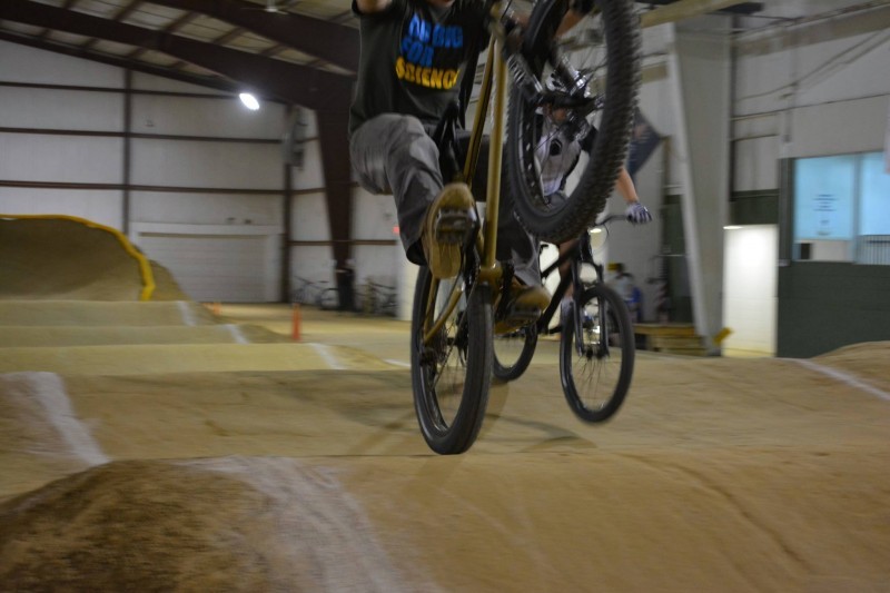 Front view of a cyclist riding a Surly Instigator bike, popping a wheelie over a whoopdeedoo, on an indoor bmx track 