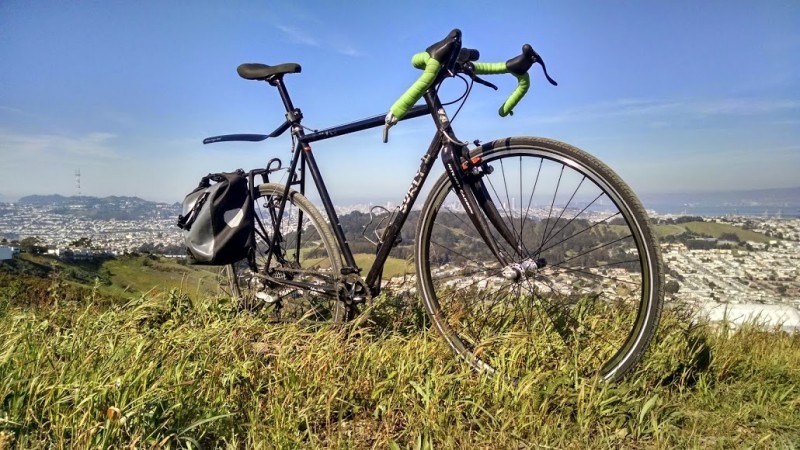 Right side view of a black Surly Cross Check bike, parked on a grassy hilltop, with hills and a city below in background