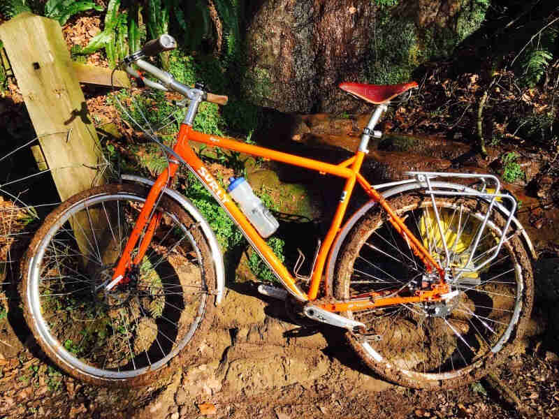 Left profile of a Surly Krampus bike, orange, parked on mud, with a forest in the background