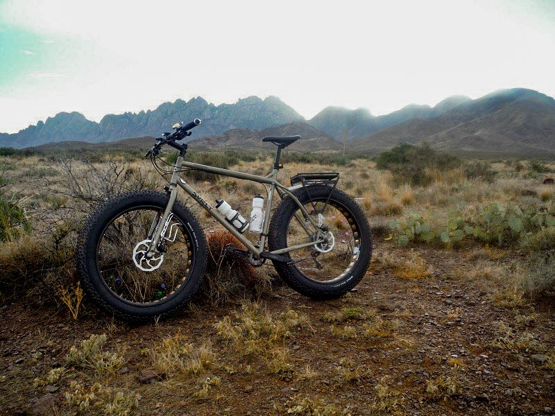 Left side view of a tan Surly Moonlander fat bike, parked in a brushy desert field, with mountains in the background