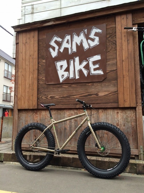 Right side view of a tan Surly Moonlander fat bike, leaning against a wood wall on a sidewalk, with a Sam's Bike sign