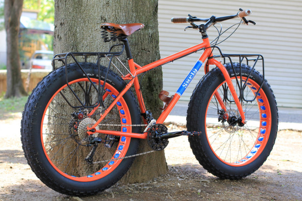 Right side view of an orange Surly Moonlander fat bike, parked in dirt against a tree, with a house wall in background