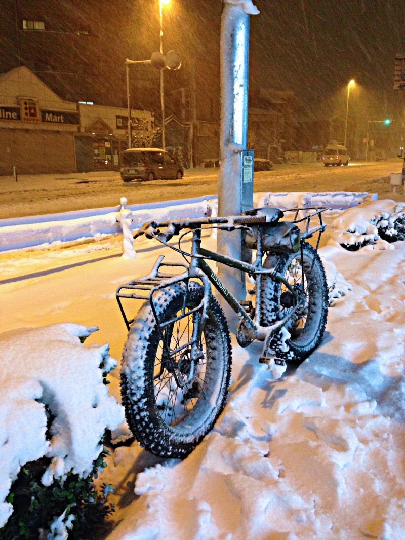 Left side view of a Surly Moonlander fat bike, on a snowy sidewalk at night