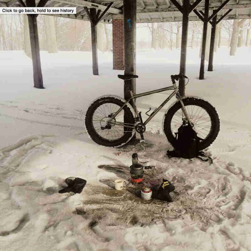 Right side view of a gray Surly Moonlander bike, parked in snow against a park shelter post, next to camp cooking items