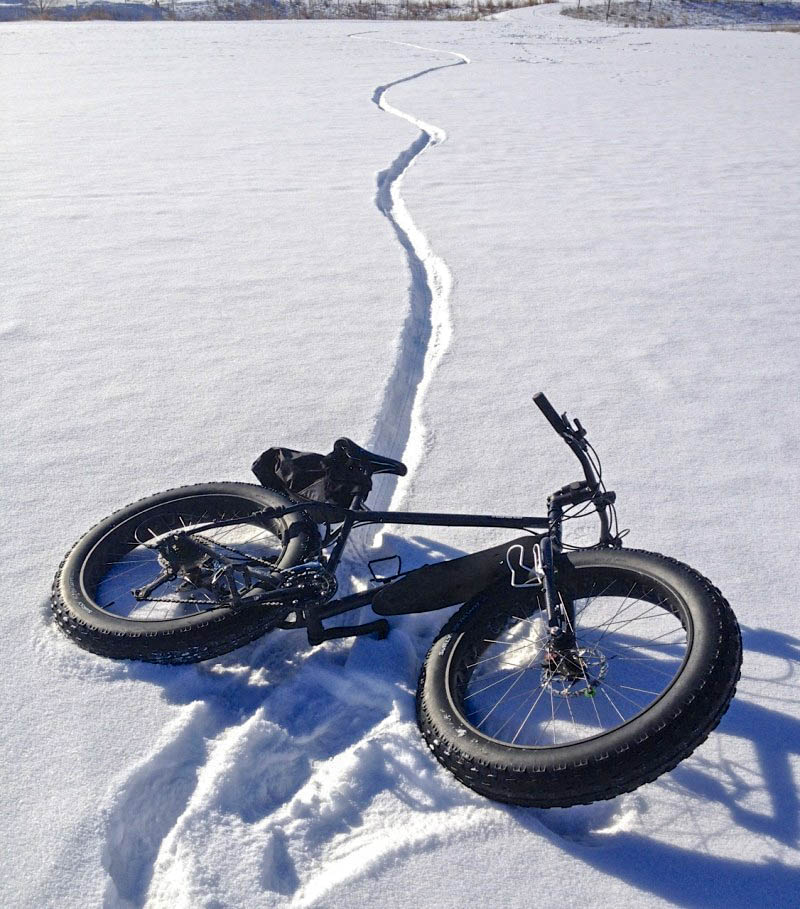 A black Surly Moonlander fat bike, laying on it's left side, on a snow covered, frozen lake, with a tire trail behind