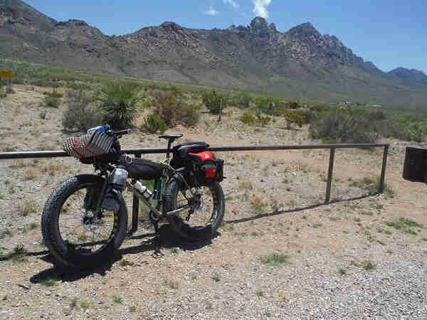 Left side view of a Surly Moonlander fat bike with gear, parked along a railing in a brushy desert with mountains behind