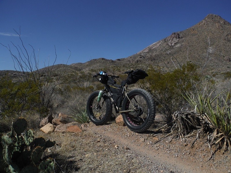 Right side view of a tan Surly Moonlander fat bike, parked on a rocky dirt trail, with desert hills in the background