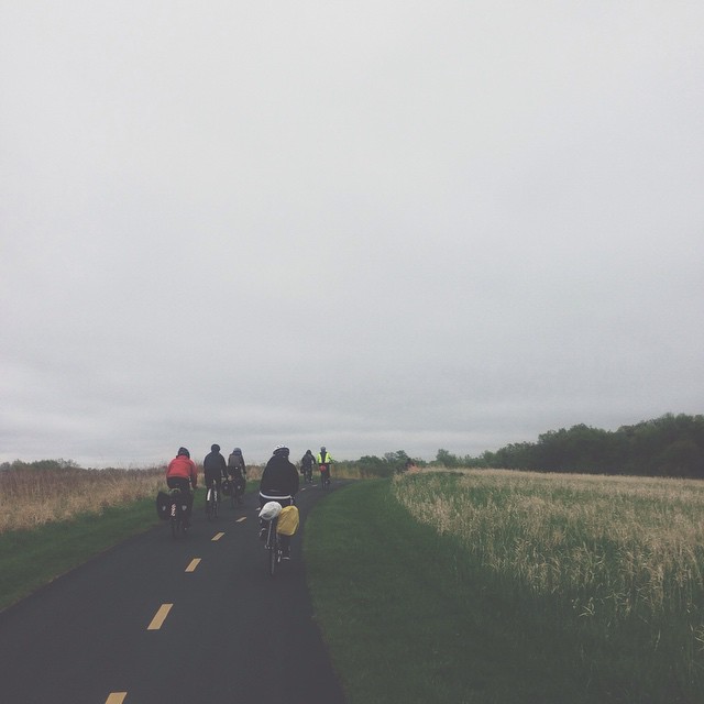 Rear view of cyclists riding down a paved bike trail running between grassy fields, on a cloudy day