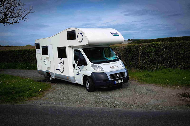 Front, right side view of a motorhome, on a gravel road, with flat topped hedge bushes behind it