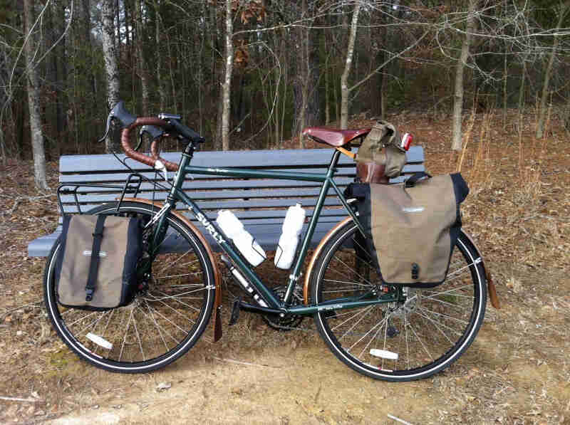 Left side view of a green Surly Disc Trucker bike with gear, parked in front of a bench, with the woods behind it
