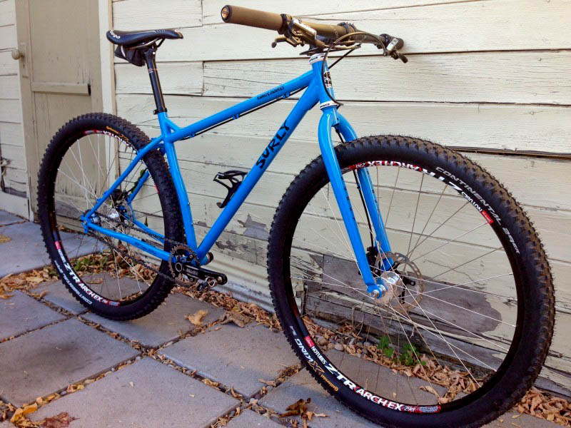 Right side view of a blue Surly Karate Monkey bike, parked on a block walkway, leaning against a wood sided wall