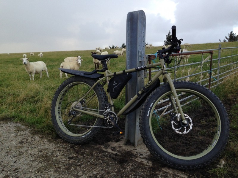 Right side view of an olive drab Surly Pugsley fat bike, leaning on a fence post with a field of sheep in the background