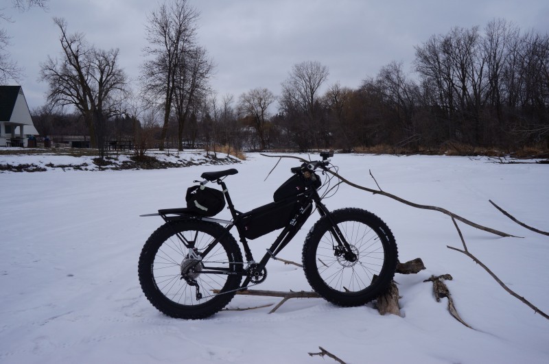 Right side view of a Surly fat bike with gear, parked on a frozen river, with bare trees and a barn in the background