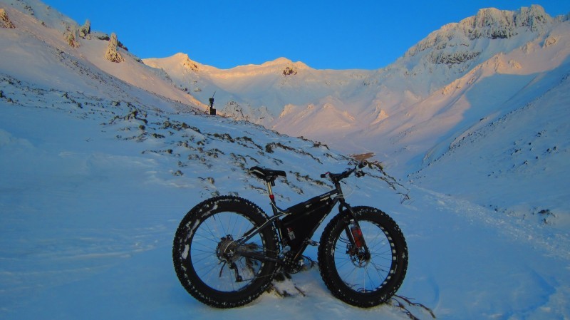 Right side view of a black Surly fat bike with frame pack, parked on a snow covered hill of a mountain 