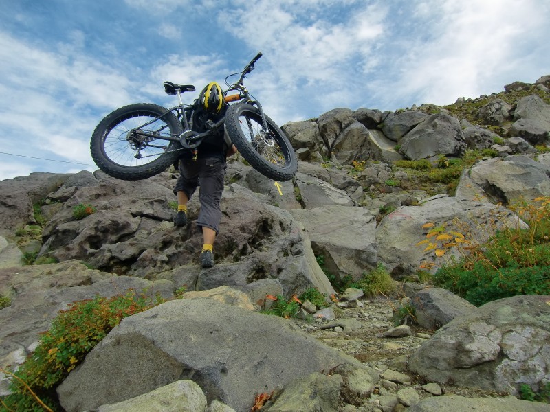 Rear, upward view of a cyclist carrying a black Surly fat bike up a steep, rock and boulder hill