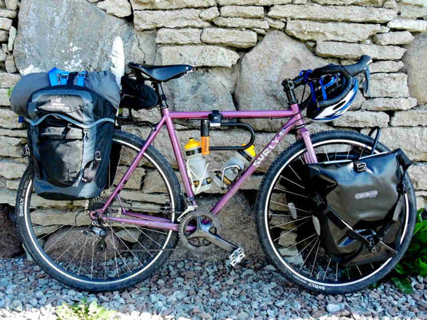 Right side view of a pink Surly bike with front and rear saddlebags, leaning against a stone wall