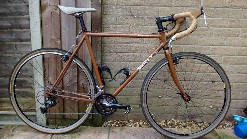 Right side view of a brown Surly Cross Check bike, leaning against a tan brick wall