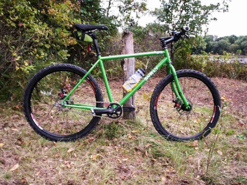Right profile view of a Surly bike, green, parked on grass in front of a wood post, with woods in the background
