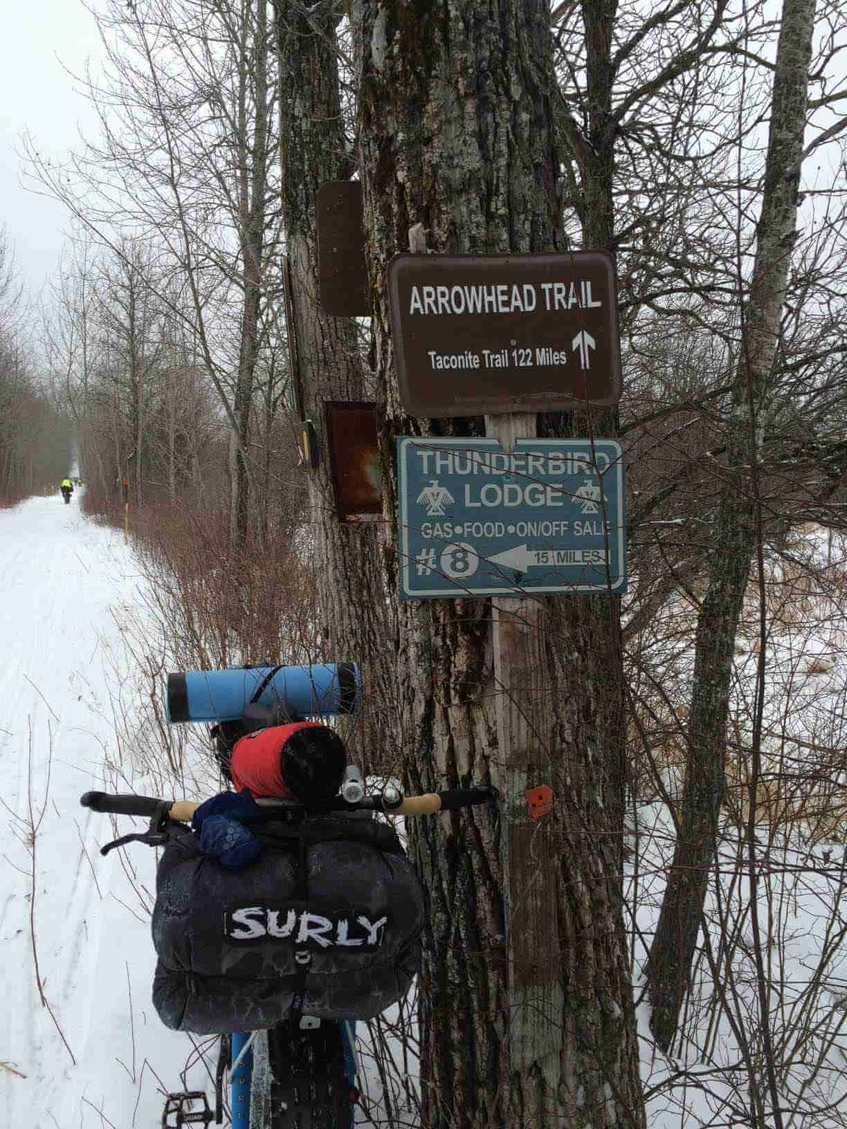 Front view of a Surly fat bike loaded with gear, parked against a tree on the side of a snowy trail