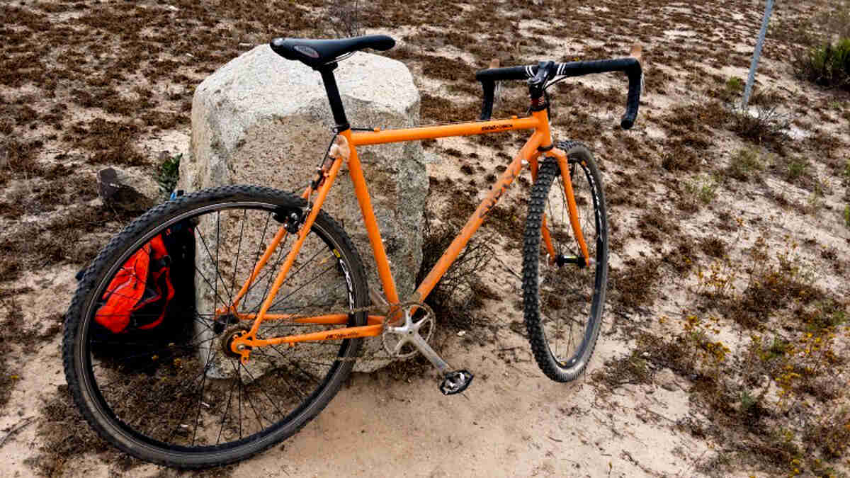 Right side view of an orange Surly bike, parked against a rock in a sandy, grass field