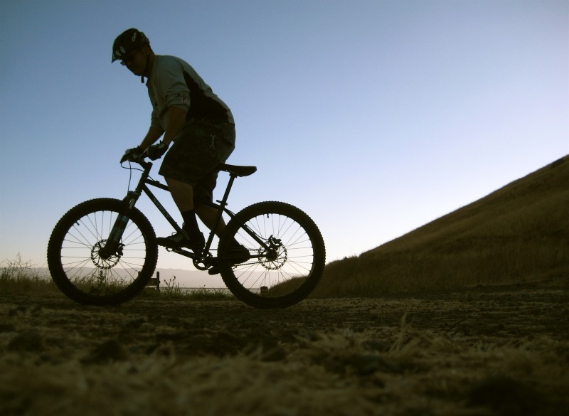 Left side view of a cyclist riding a Surly bike on a grass field with a hill in the background, in low light