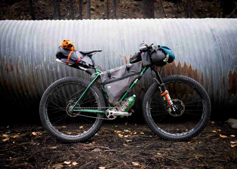 Right profile view of a green Surly bike, loaded with gear, on gravel, in front of a large corrugated tube