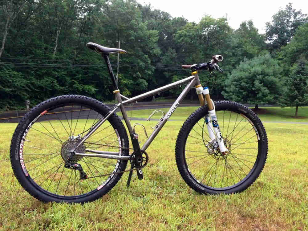 Right side view of a silver Surly bike, parked in a grass field, with a paved road and trees in the background