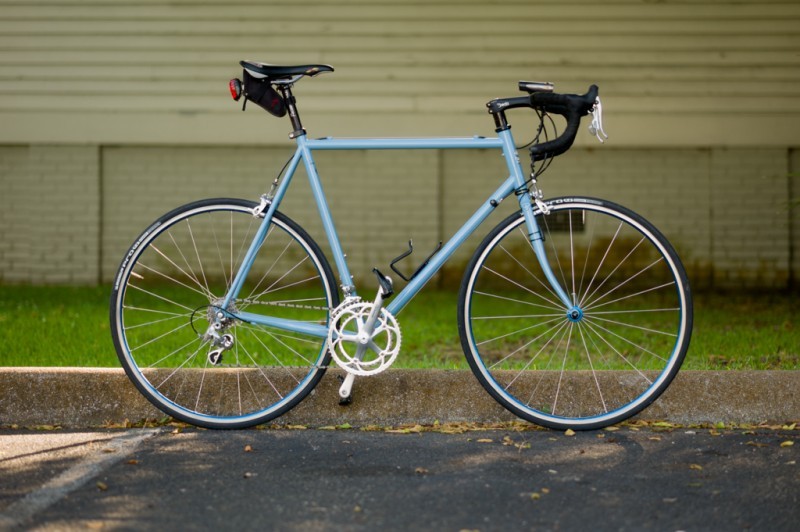 Right side view of a light blue Surly Pacer bike, parked along a curb, with a grassy patch of ground and tan wall behind