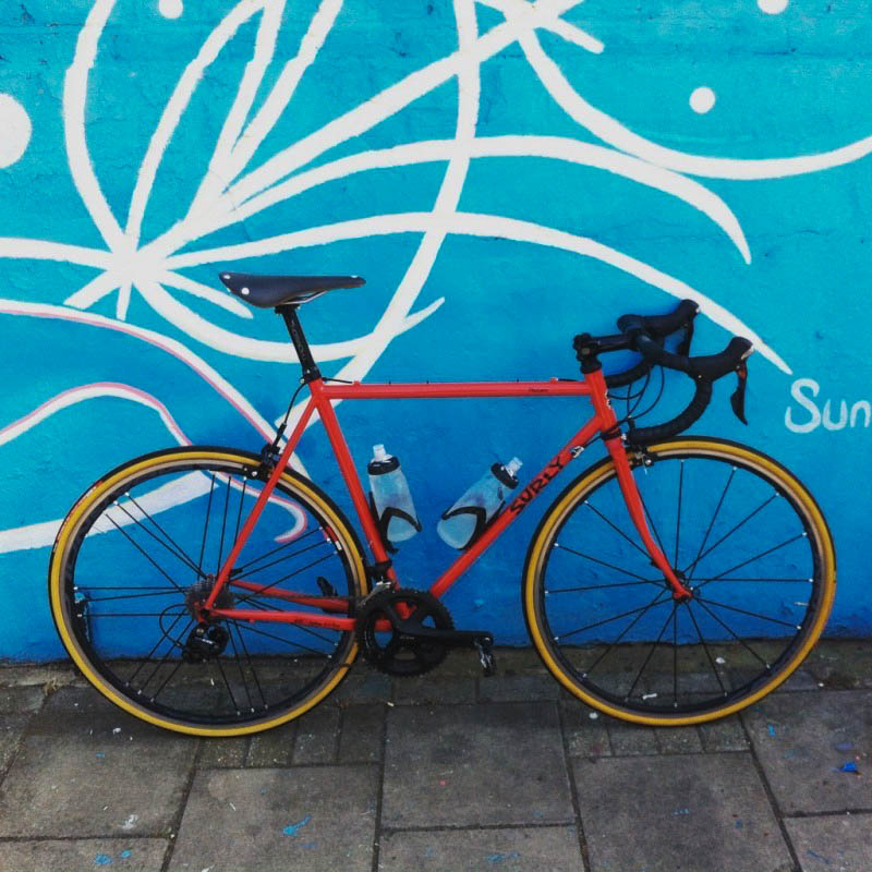 Right side view of a red Surly Pacer bike, parked on a sidewalk, leaning against a wall with a mural on it