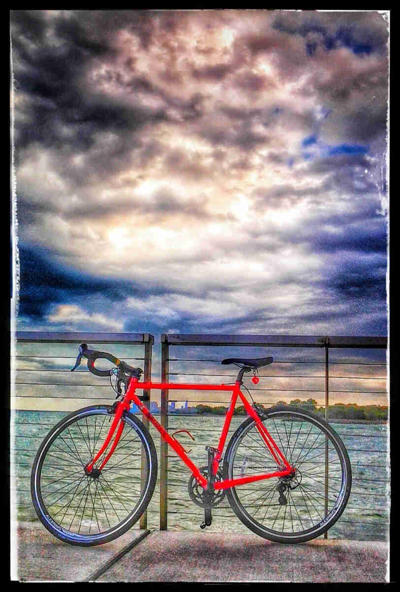 A painted portrait of a red Surly Pacer bike on a sidewalk, next to a bay,  with dark clouds above
