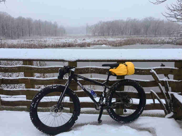 Left side view of a black Surly Ice Cream Truck fat bike, parked in the snow against a wood fence rail