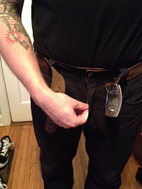 Front, chest down view of a person wearing black Surly pants, pulling the zipper up, in a room with wood floors