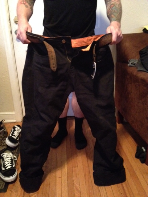Front view of a pair of black Surly pants, with a person holding them up from behind, in an interior room