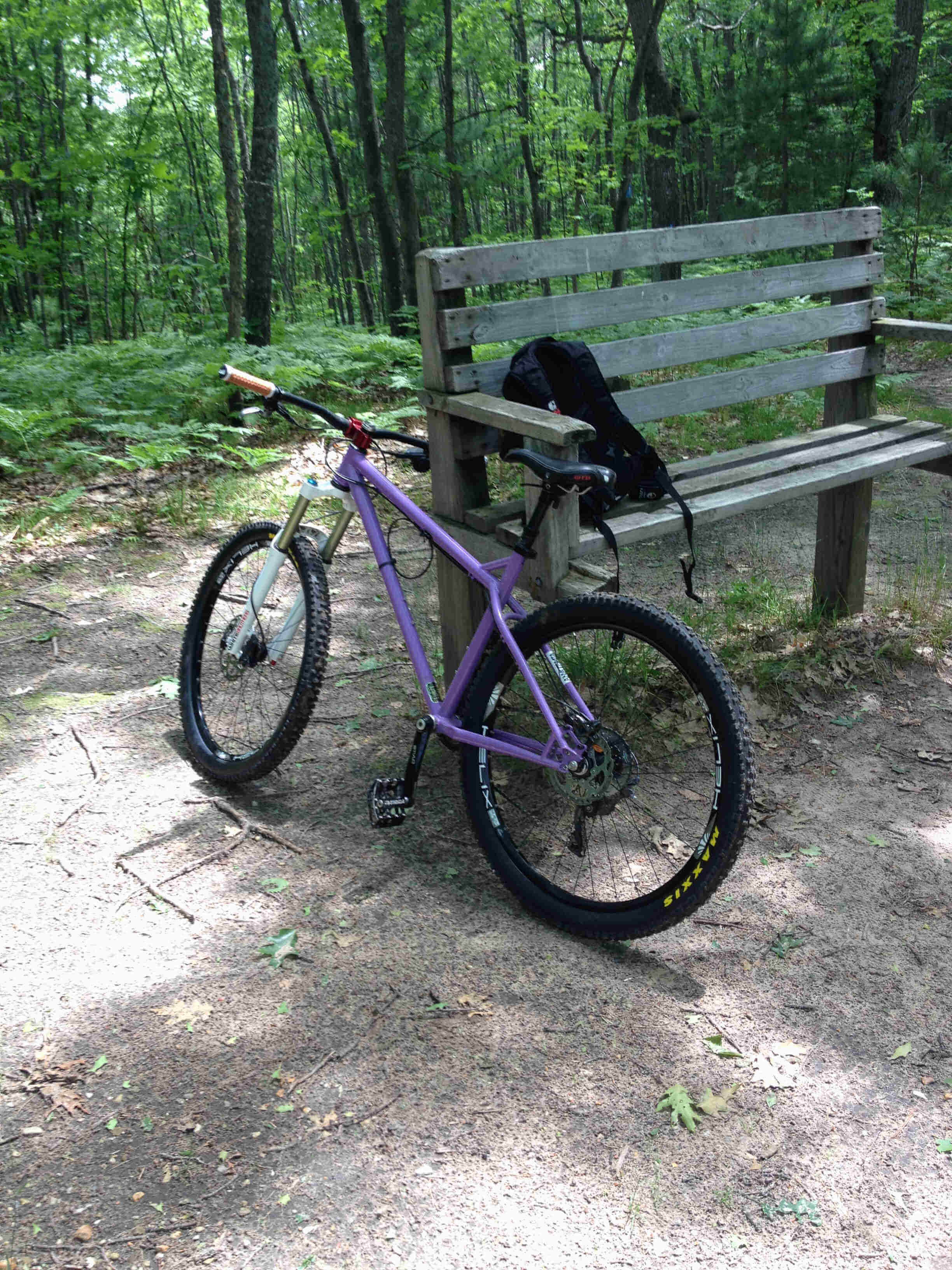 Left side view of a purple Surly Instigator bike, parked on the end a park bench, in a dirt clearing of forest