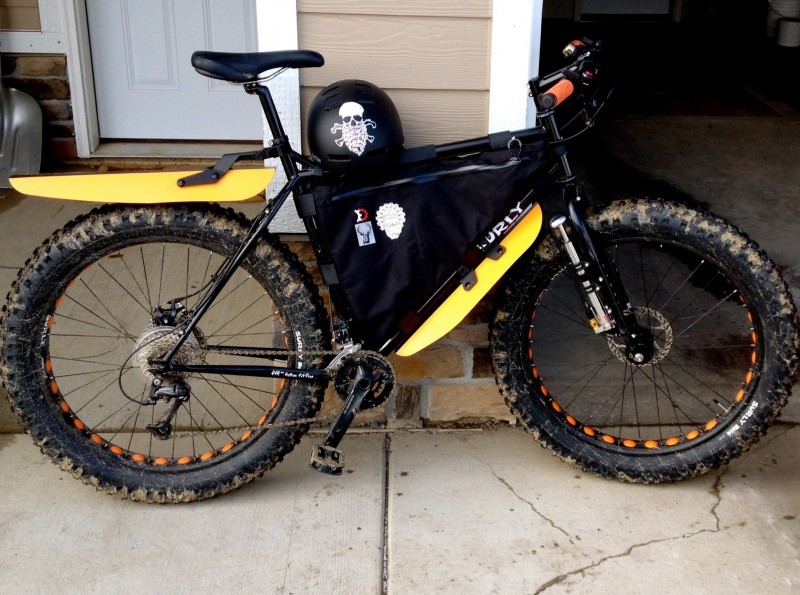 Right side view of a black Surly fat bike with a frame bag, on a walkway, leaning on a wall next to an open garage