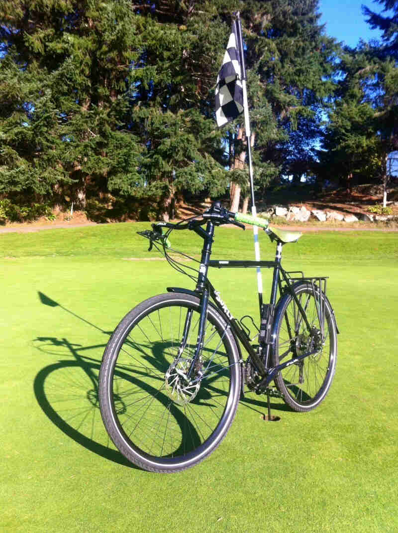 Front, left side view of a black Surly bike, leaning against the flag pin on a golf green, with pine trees in background