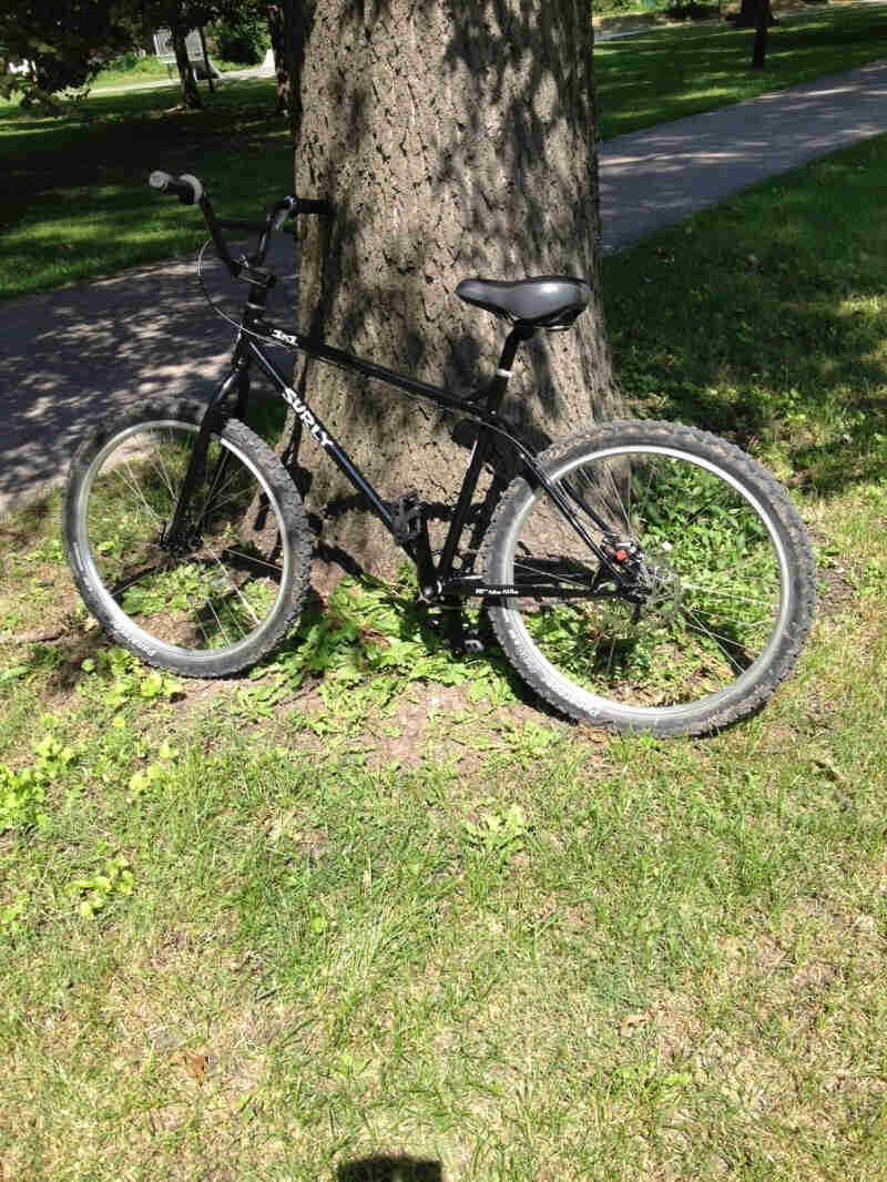 Left side view of a black Surly 1x1 bike, parked against a tree base on grass, next to a paved trail