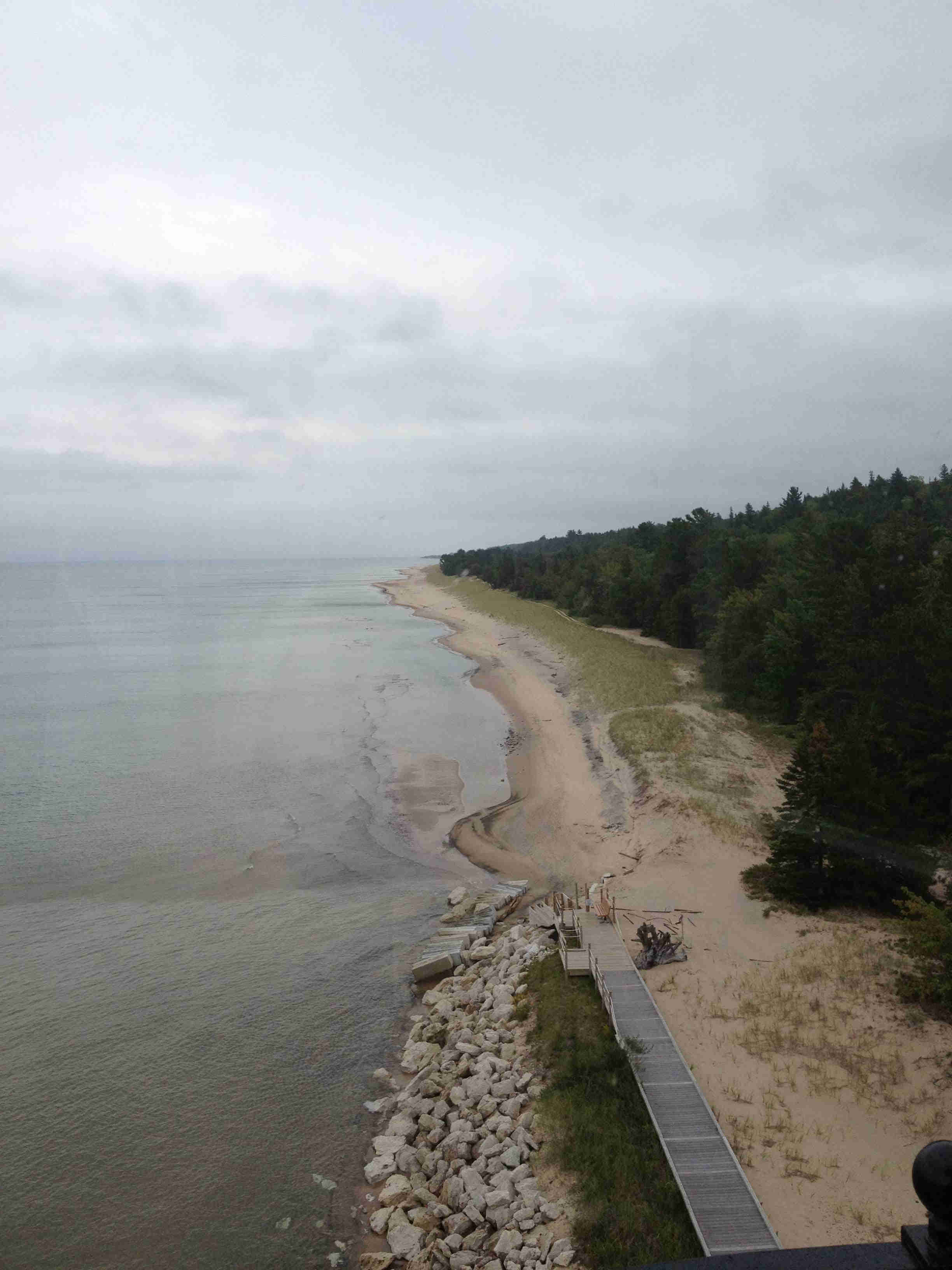 A view of above a rocky and sandy shore with a wood deck alongside, with water on the left, and trees on the right