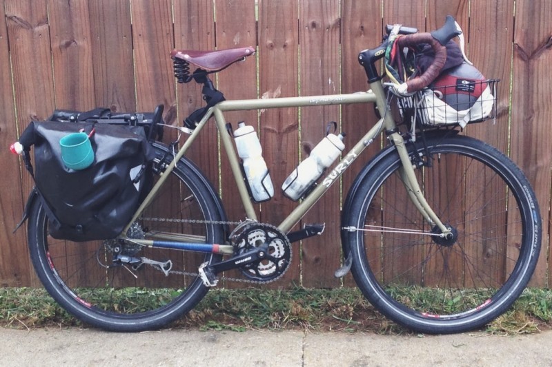 Right side view of a tan Surly Long Haul Trucker with fenders and gear, leaning against a wood fence wall, on a sidewalk