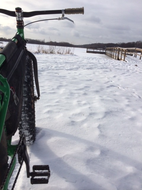 Rear, right side view of a green Surly fat bike, on a snowy field, facing a snowy frozen lake with a wood dock on it