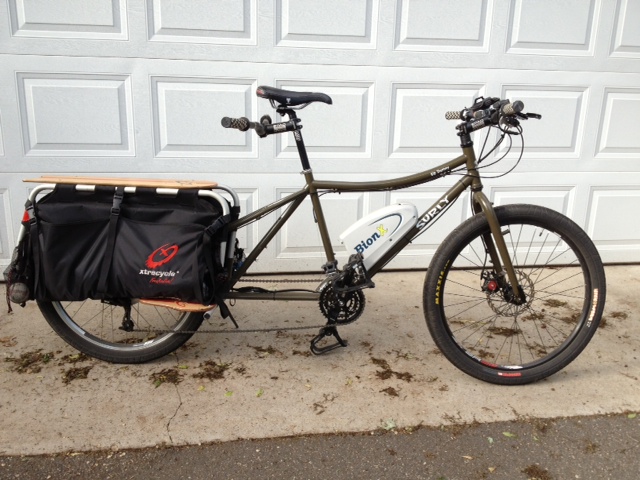 Right side view of an olive drab Surly Big Dummy bike with a BionX drive train, parked outside of a closed garage door