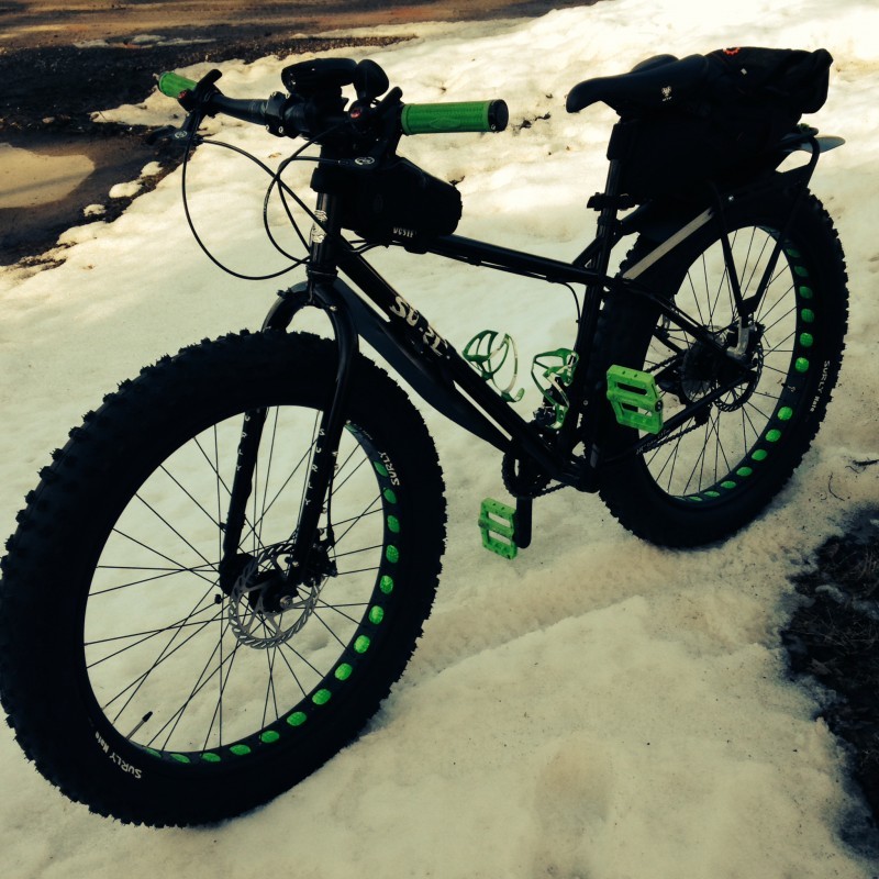 Left side view of a black Surly Pugsley fat bike, with a rear rack pack, on top of snowy ground