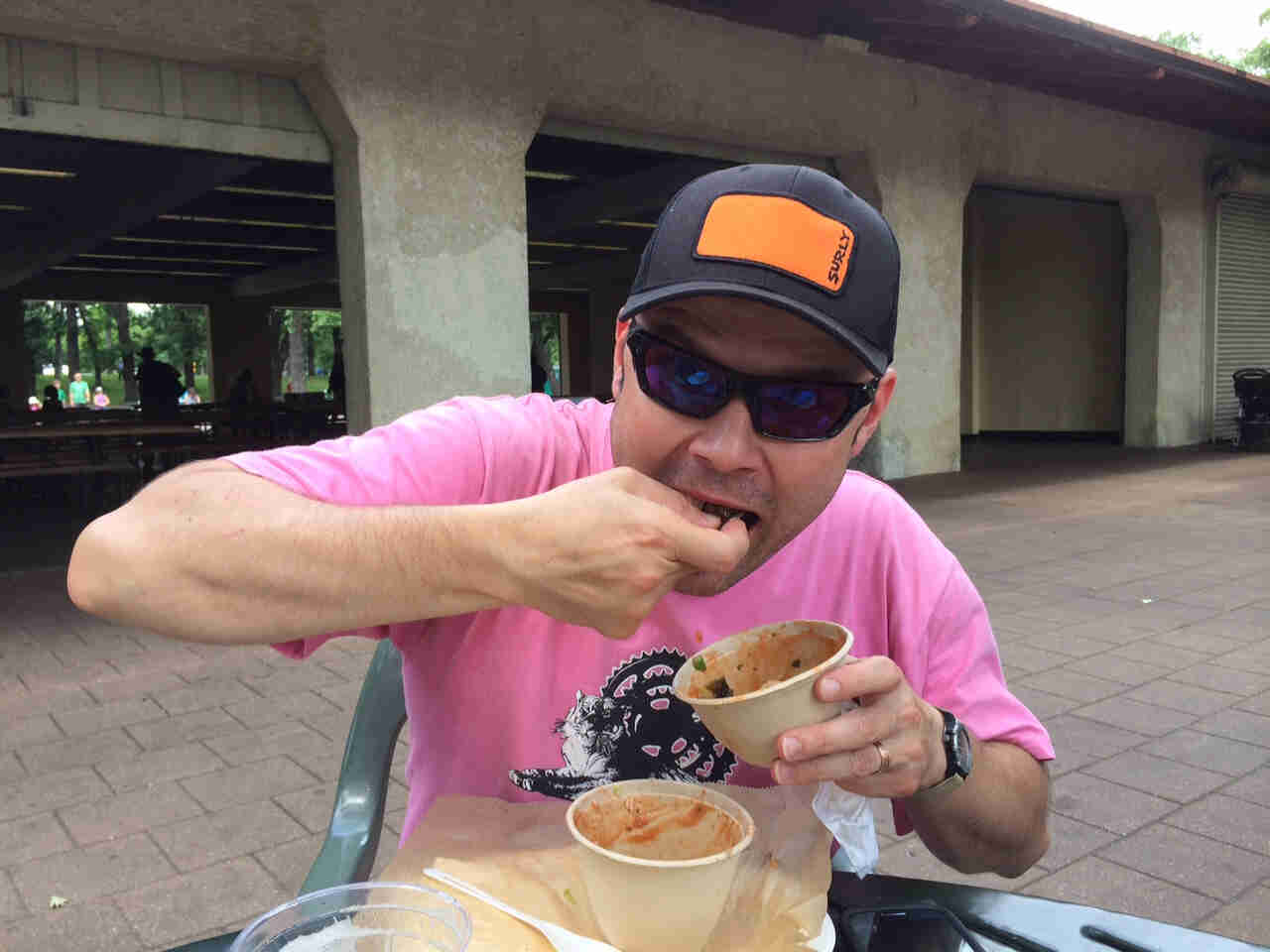 Front view of a person wearing a black Surly cap and sunglasses, sitting at a table, putting food into their mouth