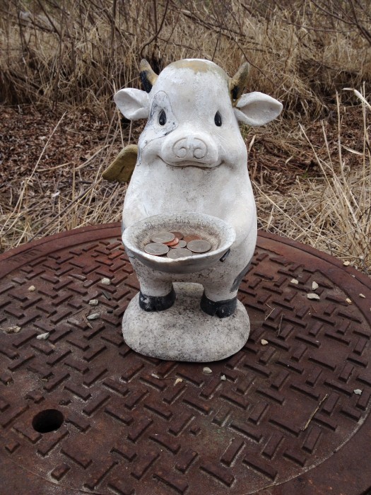 A cow figurine holding a dish with coins in it, set on a manhole cover, with a brush field behind it