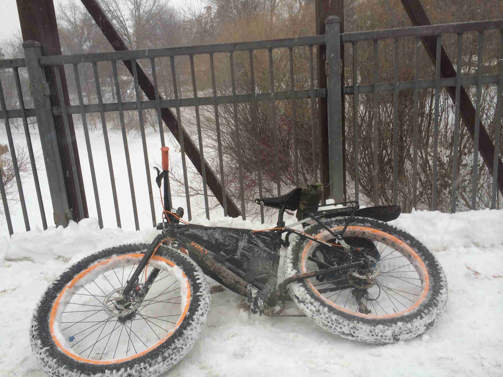Left side view of a Surly fat bike with orange rims, laying in snow, on a snow covered bridge with a frozen river below