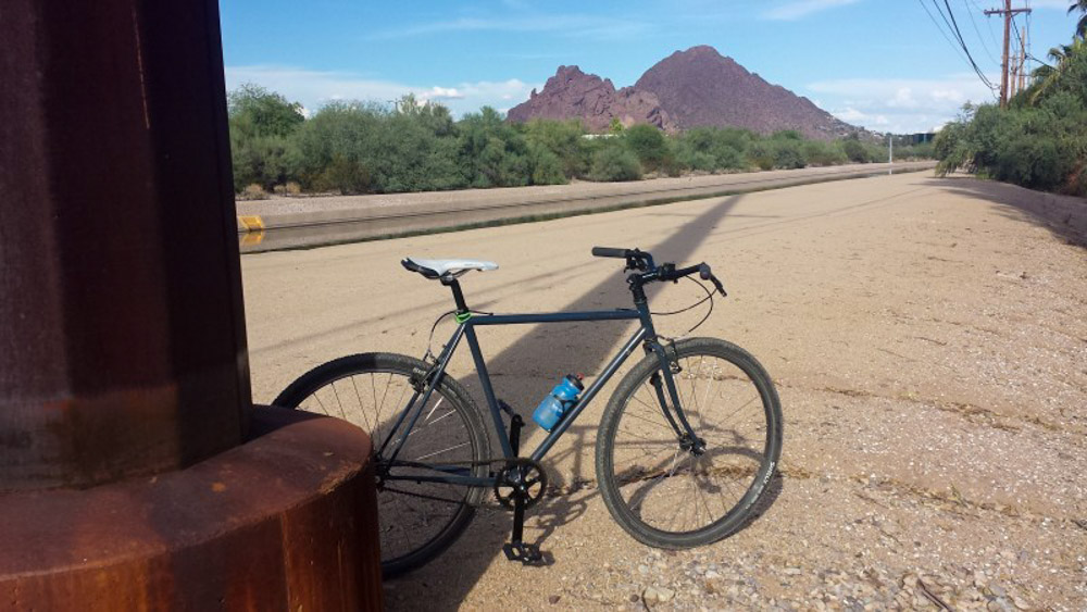 Right side view of a blue Surly Cross Check bike, parked in gravel, with bushy trees and red mountains in the background