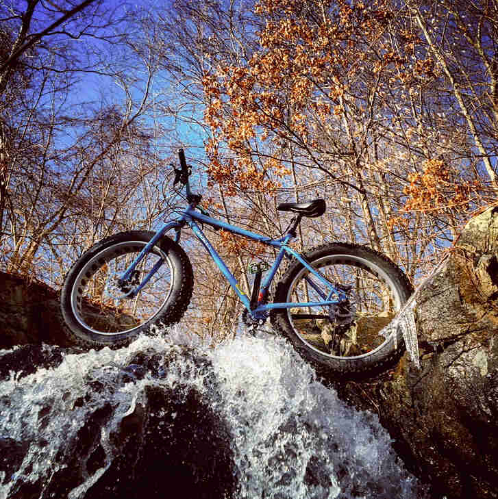 Upward, left side view of a Surly Ice Cream Truck fat bike, blue, parked across the top of a waterfall, in the woods
